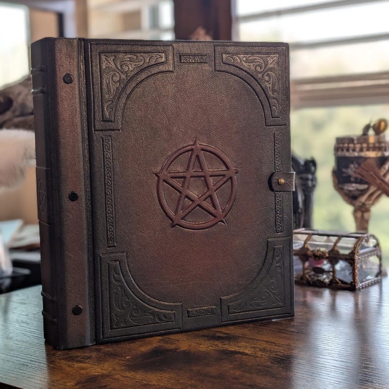 May include: A large, brown leather-bound book with a pentagram embossed on the cover. The book has decorative corner designs and a clasp closure. The book is sitting on a wooden surface.
