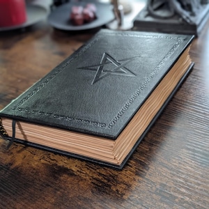 May include: A black leather-bound book with a pentagram embossed on the cover. The book has gold-colored edges and a black ribbon bookmark. The cover has decorative stitching around the edges. The book is on a dark wooden surface.