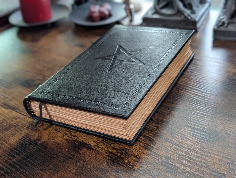 May include: A black leather-bound book with a pentagram embossed on the cover. The book has gold-colored edges and a black ribbon bookmark. The cover has decorative stitching around the edges. The book is on a dark wooden surface.