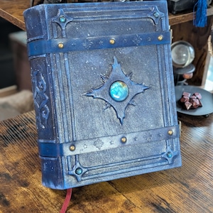 May include: A blue, fantasy-themed journal with a celestial design. The cover features a starburst emblem with a central, iridescent stone. The journal is bound with a blue strap and adorned with gold-colored rivets and decorative accents. A red ribbon bookmark is visible.