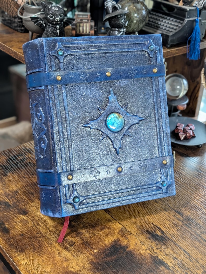 May include: A blue, fantasy-themed journal with a celestial design. The cover features a starburst emblem with a central, iridescent stone. The journal is bound with a blue strap and adorned with gold-colored rivets and decorative accents. A red ribbon bookmark is visible.