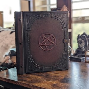 Op de afbeelding: Een donkerbruin, ledergebonden boek met een pentagram-embleem. De kaft heeft sierlijke reliëfontwerpen en een sluiting. Het boek is open en toont de rug en de pagina's.
