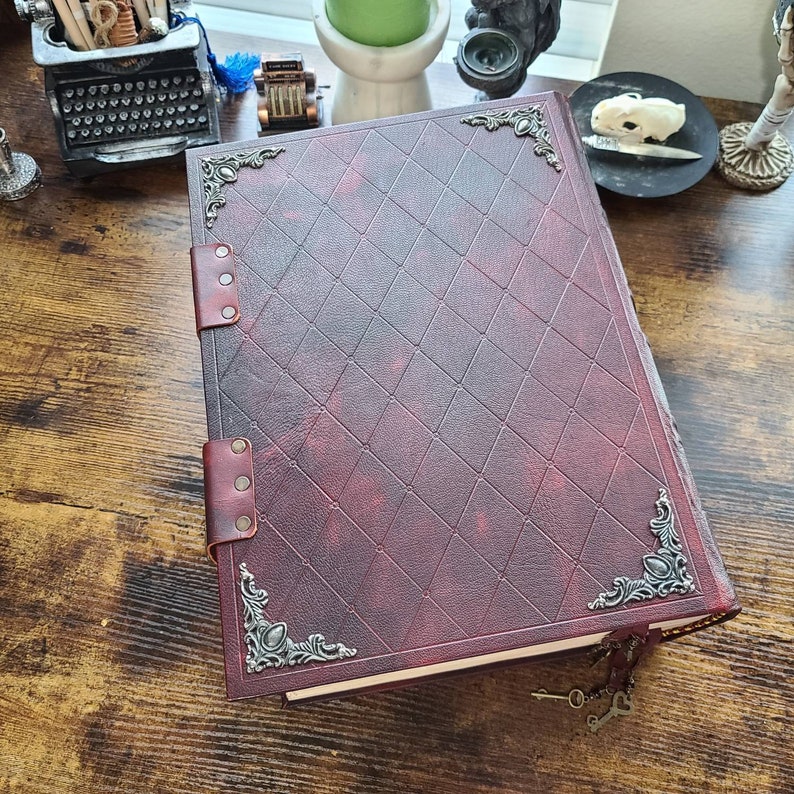 K&ouml;nnte beinhalten: Ein dunkelrotes, ledergebundenes Tagebuch mit verzierten silbernen Eckverzierungen und einem gepr&auml;gten Diamantenmuster auf dem Einband. Das Tagebuch hat zwei Lederriemen mit Metallnieten und einen Schl&uuml;sselbund, der an der Unterseite befestigt ist.
