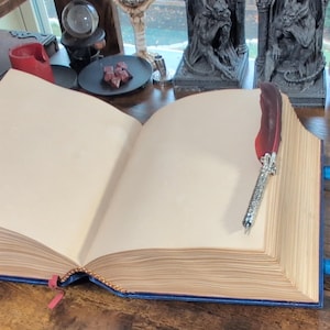 May include: An open, aged-looking book with blank pages, bound in blue leather. A decorative feather quill rests on the right page. Blue leather straps with buckles secure the book. The theme is fantasy or gothic.
