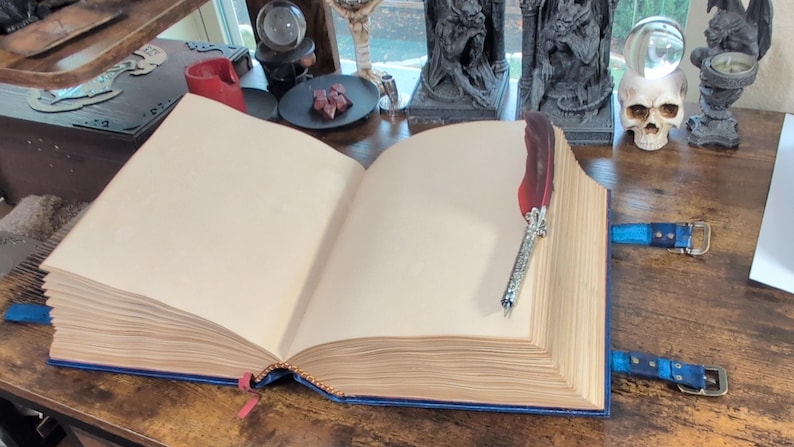 May include: An open, aged-looking book with blank pages, bound in blue leather. A decorative feather quill rests on the right page. Blue leather straps with buckles secure the book. The theme is fantasy or gothic.
