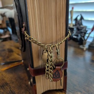 May include: A large, antique-style book with a brown leather cover and aged pages. The book is secured with a brass chain, a heart-shaped lock, and a leather key fob with a Celtic knot design. The book's spine is dark brown.