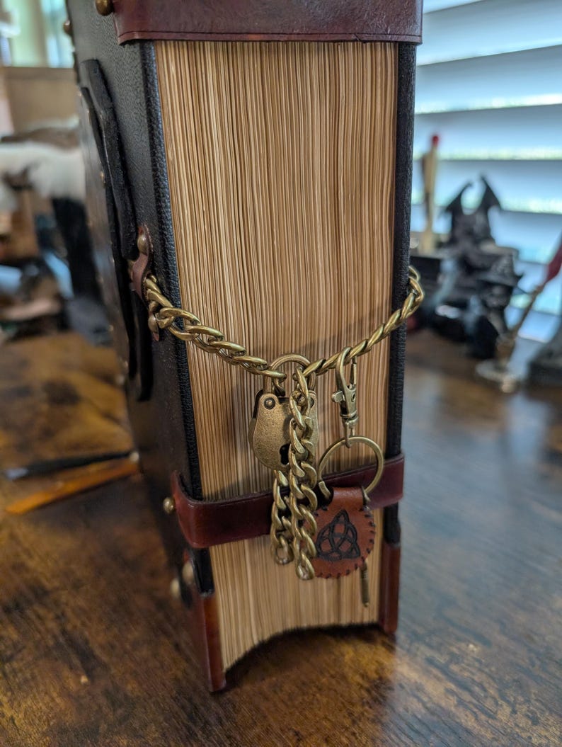 May include: A large, antique-style book with a brown leather cover and aged pages. The book is secured with a brass chain, a heart-shaped lock, and a leather key fob with a Celtic knot design. The book's spine is dark brown.