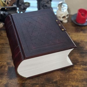 May include: A large, dark brown leather-bound book with a diamond pattern embossed on the cover. The book is closed and lying on a wooden surface.