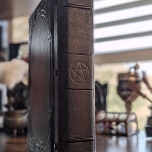 May include: A dark brown, leather-bound book with a pentagram symbol embossed on the spine. The book has decorative filigree on the front cover and is standing upright on a wooden surface. The book's spine has horizontal lines.