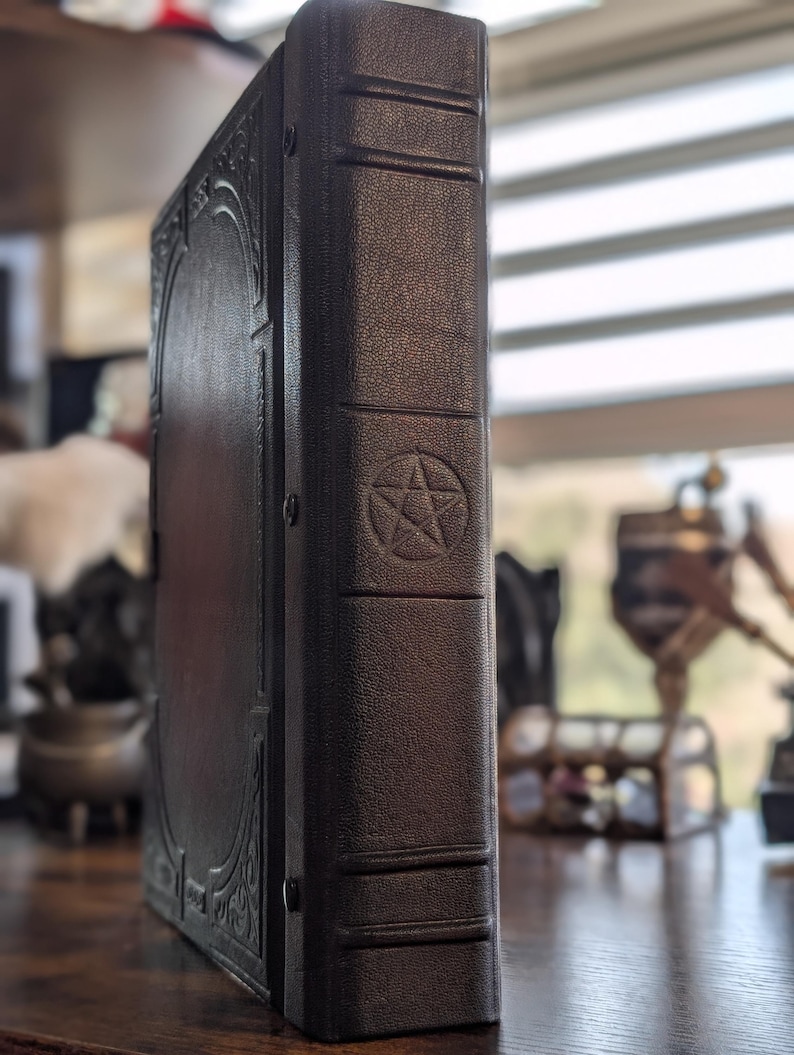 May include: A dark brown, leather-bound book with a pentagram symbol embossed on the spine. The book has decorative filigree on the front cover and is standing upright on a wooden surface. The book's spine has horizontal lines.