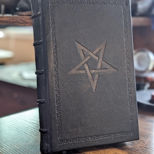 May include: A black book with a pentagram design on the cover. The book has a decorative border and a ribbon bookmark. The spine has raised bands. The book is standing upright on a wooden surface.