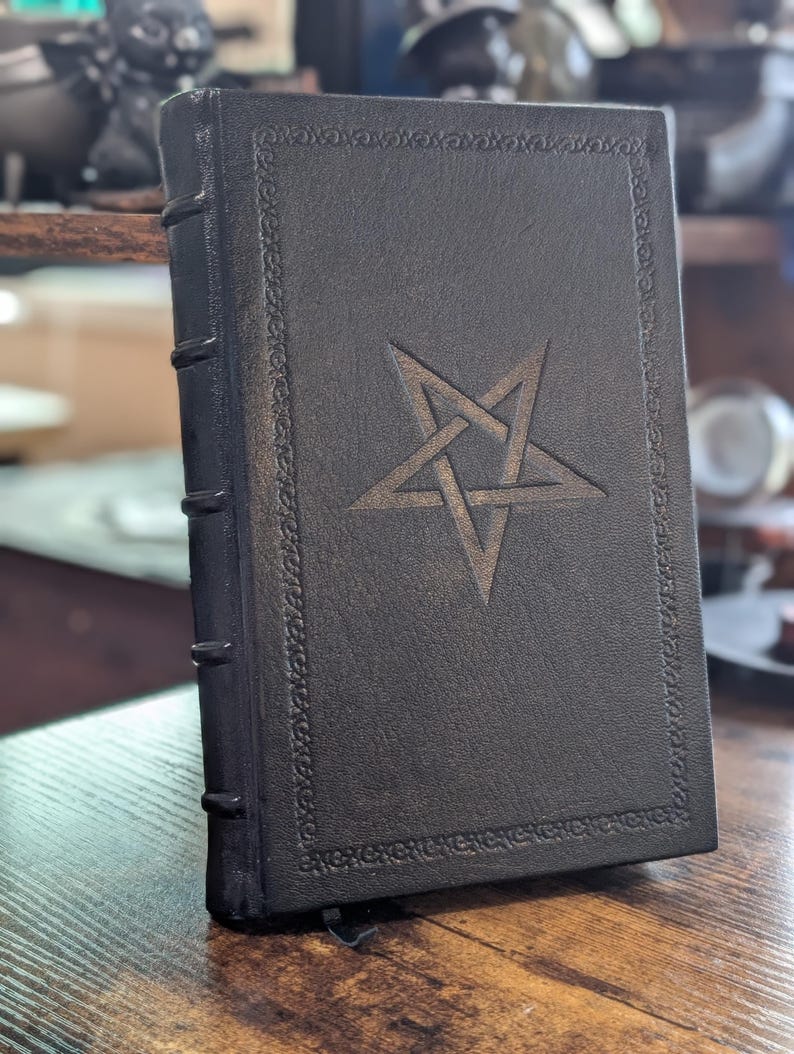 May include: A black book with a pentagram design on the cover. The book has a decorative border and a ribbon bookmark. The spine has raised bands. The book is standing upright on a wooden surface.