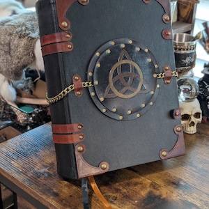 May include: A large, black book with brown leather accents and brass hardware. The cover features a circular emblem with a gold Celtic knot design, secured by a gold chain. A brown ribbon bookmark hangs from the pages. The book is on a wooden surface.