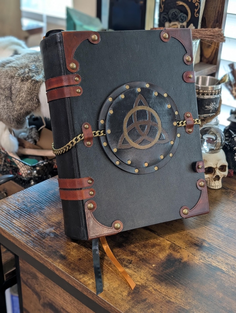 May include: A large, black book with brown leather accents and brass hardware. The cover features a circular emblem with a gold Celtic knot design, secured by a gold chain. A brown ribbon bookmark hangs from the pages. The book is on a wooden surface.