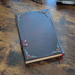 Puede incluir: Un diario de cuero burdeos oscuro con adornos de esquina plateados ornamentados y adornos de piedras preciosas rojas. El diario tiene un cierre de correa de cuero y un marcador de cinta roja. El cuaderno est&aacute; sobre una superficie de madera.