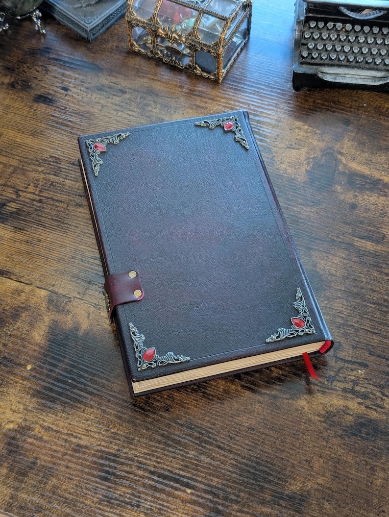 Puede incluir: Un diario de cuero burdeos oscuro con adornos de esquina plateados ornamentados y adornos de piedras preciosas rojas. El diario tiene un cierre de correa de cuero y un marcador de cinta roja. El cuaderno est&aacute; sobre una superficie de madera.