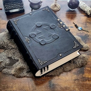 May include: A black leather-bound journal with a decorative metal clasp and a raised design of a circle with five points on the front cover. The journal is lying on a brown wooden surface with a fur rug underneath.