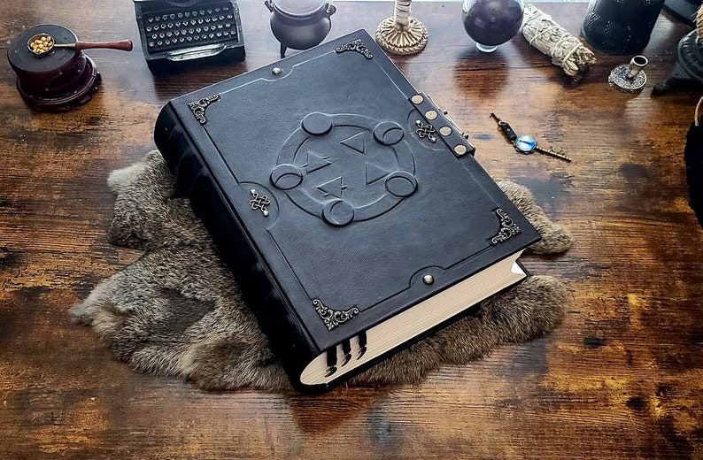 May include: A black leather-bound journal with a decorative metal clasp and a raised design of a circle with five points on the front cover. The journal is lying on a brown wooden surface with a fur rug underneath.