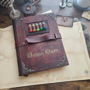 May include: A brown leather-bound journal with a metal clasp and decorative accents. The journal has the words "Dungeon Master" embossed on the front cover. There are five small glass vials with colorful liquids inside, attached to the front cover.