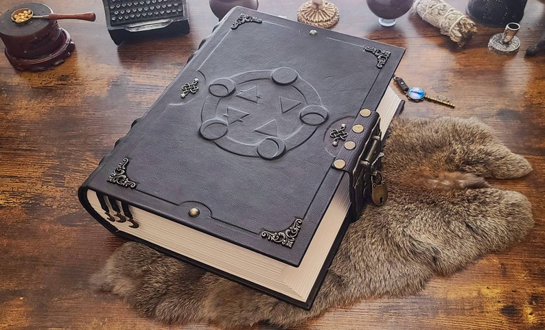 May include: A large, black leather-bound journal with a decorative lock and key. The cover features an embossed design of a pentagram with four moons around it.