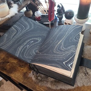 May include: A large, open journal with black leather binding and marbled pages. The pages feature a swirling white and gold pattern. The journal is on a wooden surface, with a decorative clasp and ribbon bookmark.