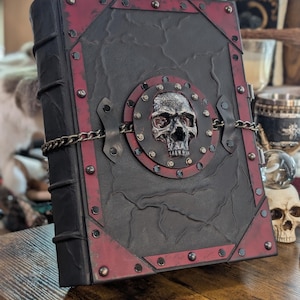 May include: A large, gothic-style book with a black cover and red trim. A metal skull is centered on the front, surrounded by a circular plate and chain. The book has metal corner protectors and a textured surface.