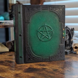 Op de afbeelding: Een rechthoekig boek met een donkergroene leren kaft met een in reliëf aangebracht pentagram in het midden. De kaft heeft een donkere, verweerde rand met ingewikkelde krulpatronen en een rug die is vastgezet met drie zwarte klinknagels.