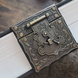 May include: A black leather-bound journal with a decorative metal clasp. The clasp features an ornate design with a keyhole and a small metal handle.