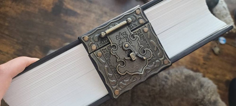 May include: A black leather-bound journal with a decorative metal clasp. The clasp features an ornate design with a keyhole and a small metal handle.