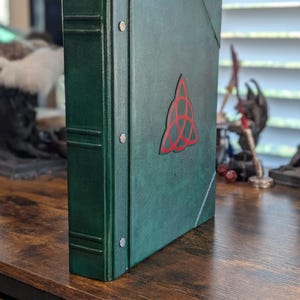 May include: A dark green binder with a red Celtic knot design on the front. The binder has a textured appearance and metal rivets along the spine. The binder is standing upright on a wooden surface.