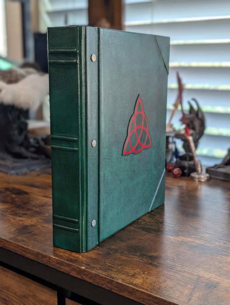 May include: A dark green binder with a red Celtic knot design on the front. The binder has a textured appearance and metal rivets along the spine. The binder is standing upright on a wooden surface.