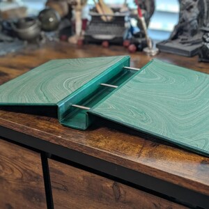 May include: A green book stand with a marbled pattern. The stand is open, with two angled surfaces held together by metal rods. The stand is on a wooden surface.