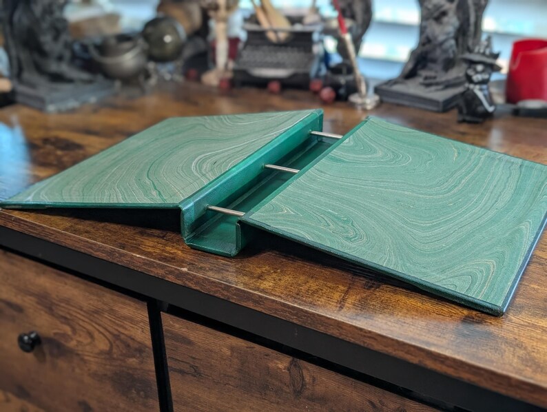 May include: A green book stand with a marbled pattern. The stand is open, with two angled surfaces held together by metal rods. The stand is on a wooden surface.