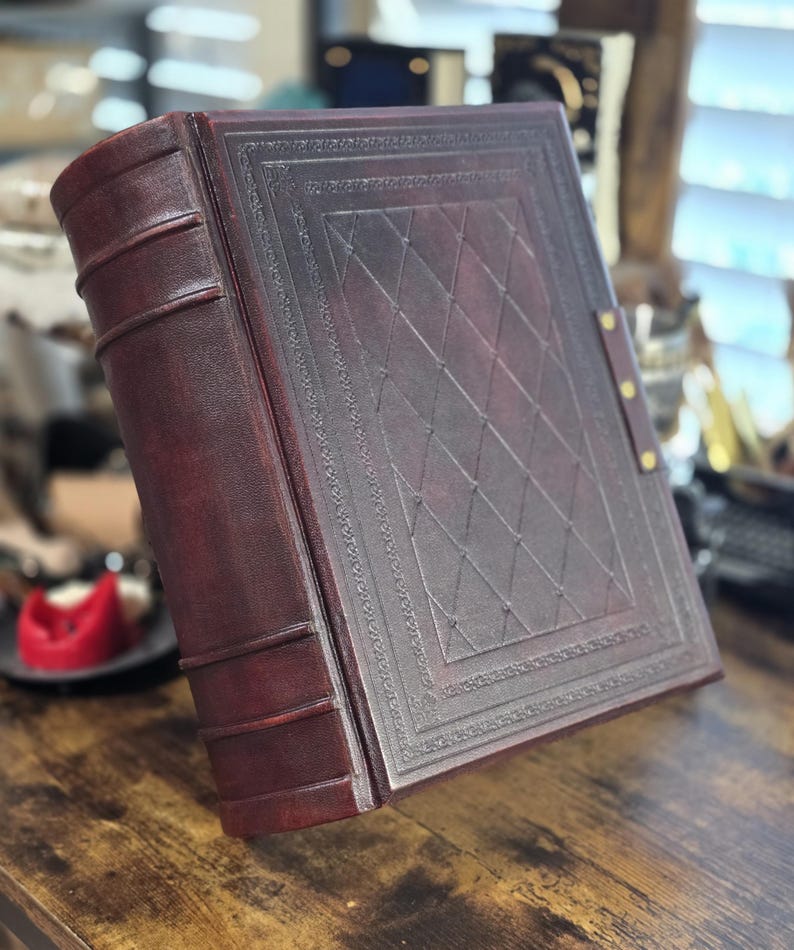 May include: A dark brown leather-bound journal with a decorative embossed pattern and brass accents. The journal has a spine with raised ridges and a decorative border around the cover.