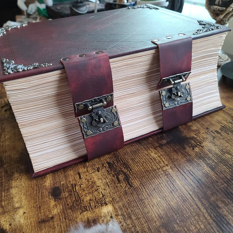 K&ouml;nnte beinhalten: Ein gro&szlig;es, dickes, ledergebundenes Buch mit zwei Metallschlie&szlig;en und dekorativen Metallakzenten. Das Buch ist geschlossen und liegt auf einer Holzoberfl&auml;che.