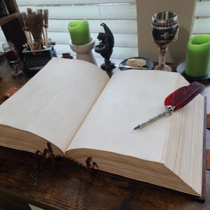 K&ouml;nnte beinhalten: Ein offenes, leeres, ledergebundenes Tagebuch mit einer roten Federkielstift, die auf den ge&ouml;ffneten Seiten liegt. Das Tagebuch liegt auf einer Holzoberfl&auml;che mit anderen Dekorationsgegenst&auml;nden, darunter Kerzen und ein Metallkelch.