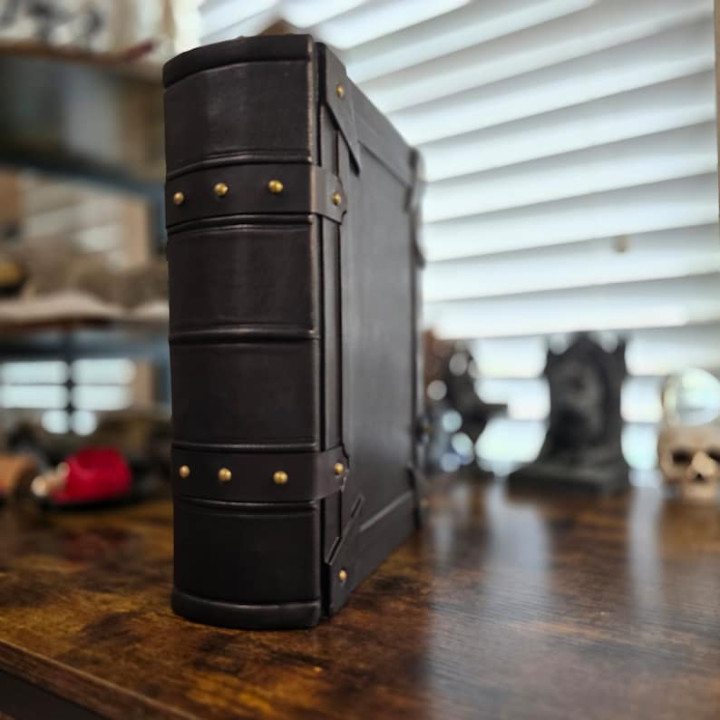 May include: A black leather-bound journal with brass accents and metal clasps. The journal is closed and standing upright on a wooden surface.