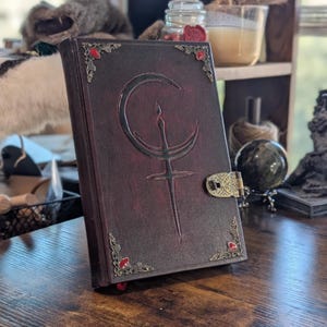 Puede incluir: Un diario de cuero burdeos con un dise&ntilde;o de luna creciente y espada en relieve en la portada. El diario presenta detalles decorativos en las esquinas con gemas rojas y un cierre de lat&oacute;n. Las p&aacute;ginas se cierran con una cinta roja.