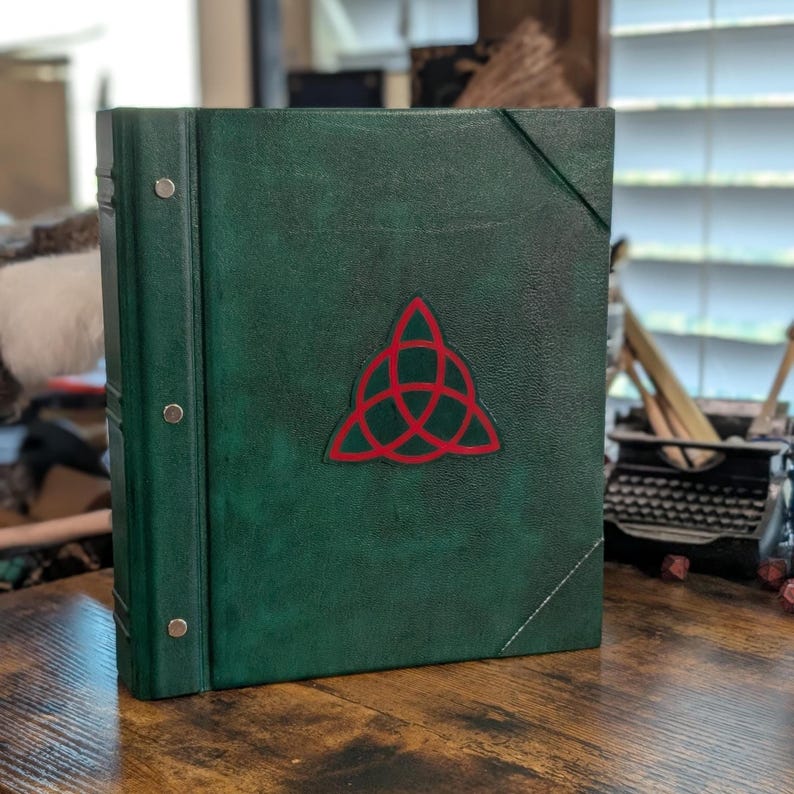 May include: A dark green leather-bound book with a red Celtic knot emblem. The book has metal ring binders and a strap closure. The cover is textured, and the book is on a wooden surface.