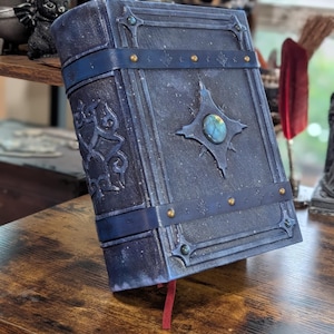 May include: A dark blue, faux leather journal with a gemstone centerpiece and decorative metal accents. The journal has a dark blue strap and gold-colored rivets. A red ribbon bookmark is visible.