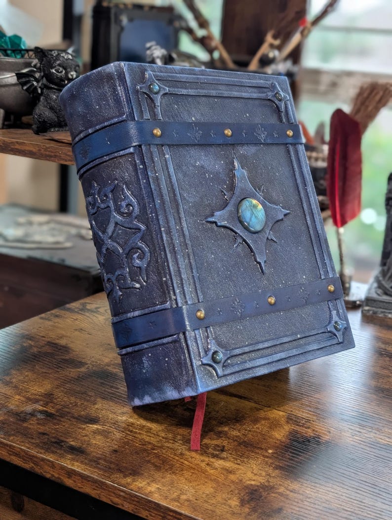 May include: A dark blue, faux leather journal with a gemstone centerpiece and decorative metal accents. The journal has a dark blue strap and gold-colored rivets. A red ribbon bookmark is visible.