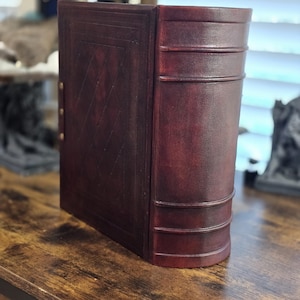 May include: A burgundy leather-bound book with a decorative embossed pattern on the front cover. The book has a spine with three raised bands and is closed.