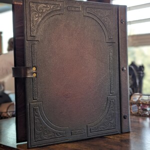 May include: A dark brown leather-bound journal with an embossed design. The cover features an arched border with ornate corner details and a metal clasp. The journal is open, revealing the interior pages, and rests on a wooden surface.
