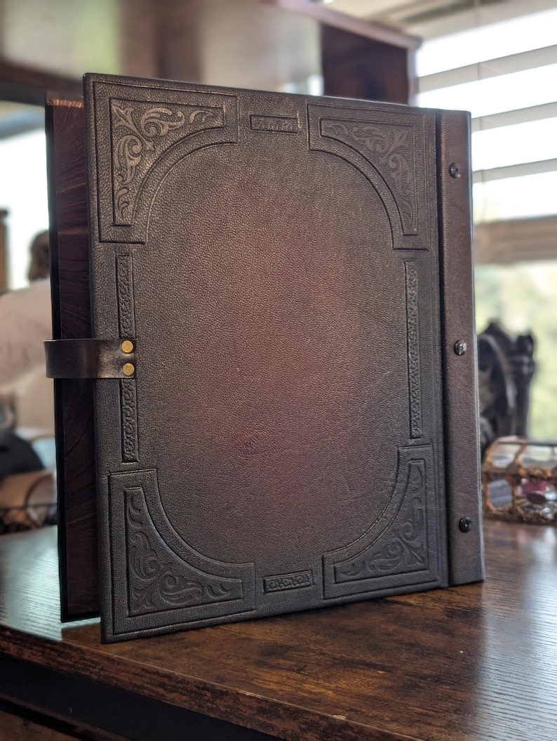 May include: A dark brown leather-bound journal with an embossed design. The cover features an arched border with ornate corner details and a metal clasp. The journal is open, revealing the interior pages, and rests on a wooden surface.
