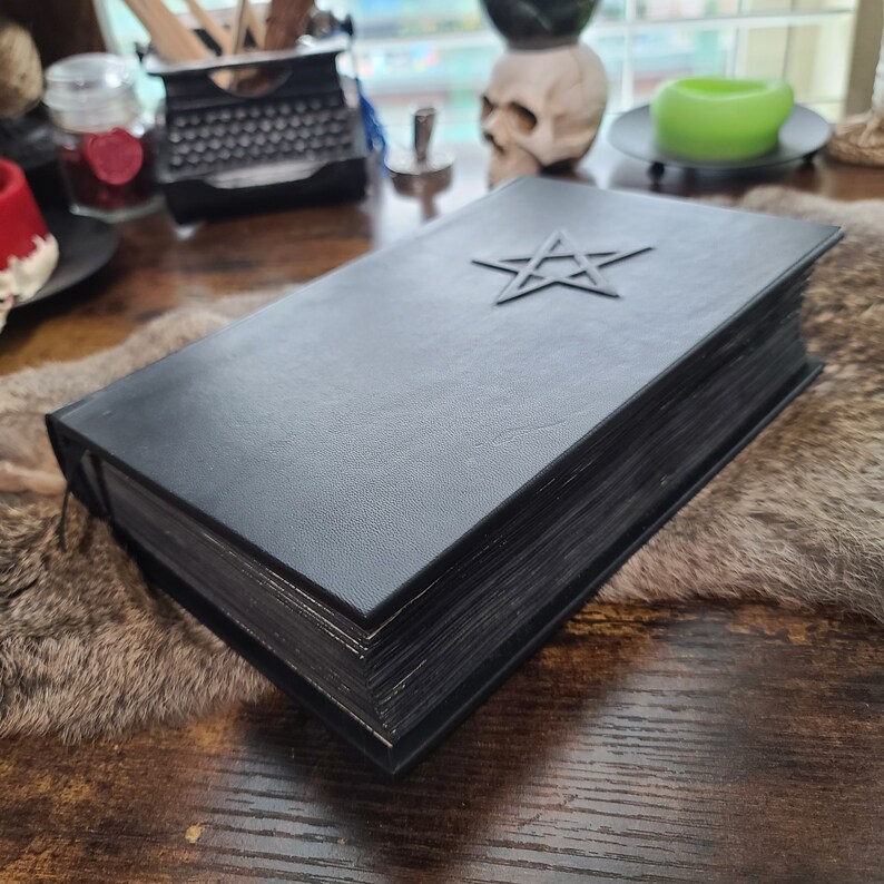 May include: A black leather-bound book with a raised pentagram on the cover. The book has thick, dark-edged pages and a black ribbon bookmark. The book is on a textured surface.