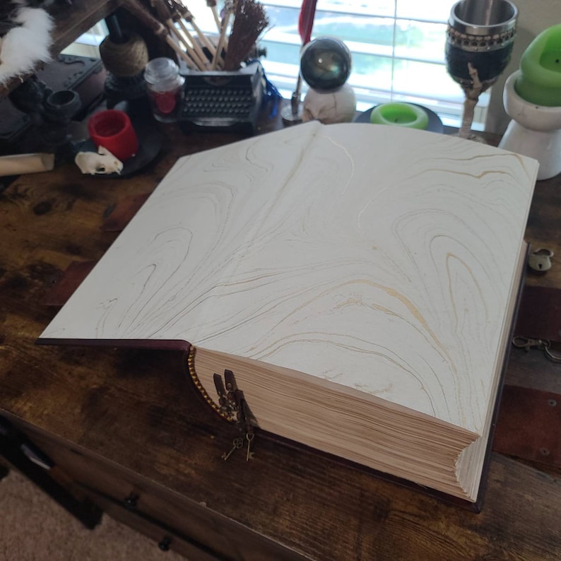 K&ouml;nnte beinhalten: Ein gro&szlig;es, antik aussehendes Buch mit einem burgunderfarbenen Lederbezug und goldenen Akzenten. Das Buch ist ge&ouml;ffnet und zeigt wei&szlig;e Seiten mit goldenen Marmorierungen. Das Buch ist mit einem Schl&uuml;sselsatz verschlossen.