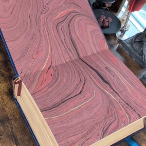 May include: Open book with marbled pink, black, and gold patterned pages. The book has a blue cover and a red ribbon bookmark. Blue leather straps with buckles are attached to the cover.