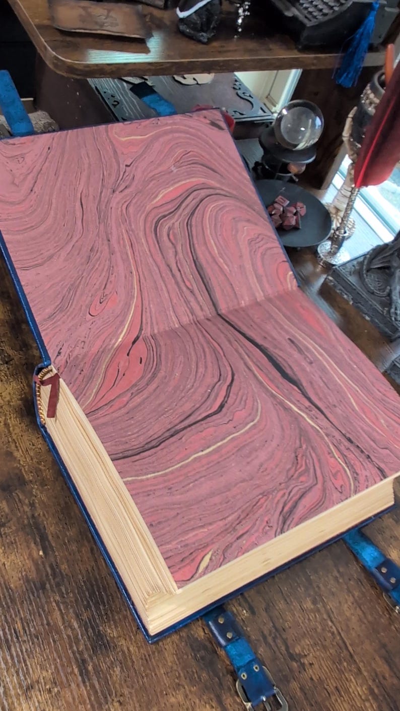 May include: Open book with marbled pink, black, and gold patterned pages. The book has a blue cover and a red ribbon bookmark. Blue leather straps with buckles are attached to the cover.