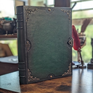May include: A dark green leather-bound journal with ornate corner and clasp details. The journal has a textured cover and a spine with raised bands. Decorative metal accents adorn the corners and clasp, adding a vintage aesthetic. The journal sits on a wooden surface.