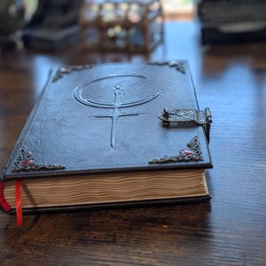 Puede incluir: Un diario de cuero marr&oacute;n oscuro con un cierre decorativo y adornos en las esquinas. La cubierta presenta un dise&ntilde;o en relieve de una luna creciente y una espada. Una cinta roja de marcador es visible, y las p&aacute;ginas tienen un aspecto envejecido.